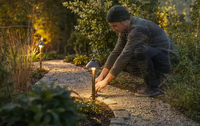 Person richtet eine niedrige warm leuchtende Gartenlampe am Kiesweg aus; Thema Einbaupools, Schwerpunkt Reparatur; Ergebnis.