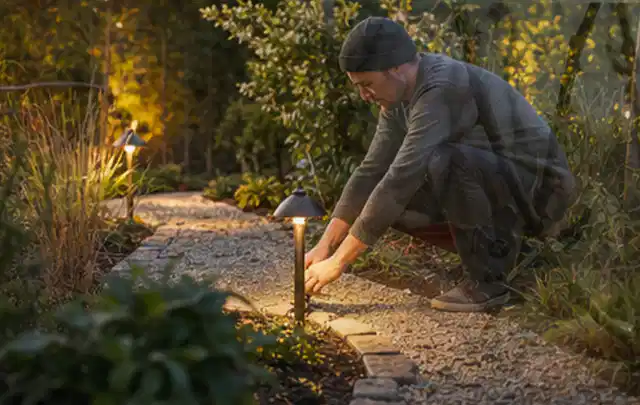 Person richtet eine niedrige warm leuchtende Gartenlampe am Kiesweg aus; Thema Einbaupools, Schwerpunkt Sicherheit; Praxisalltag.