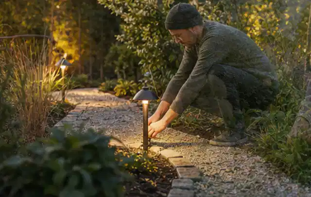 Person richtet eine niedrige warm leuchtende Gartenlampe am Kiesweg aus; Thema Einbaupools, Schwerpunkt Vergleich; Ergebnis.