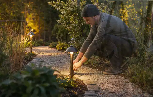 Person richtet eine niedrige warm leuchtende Gartenlampe am Kiesweg aus; Thema Familiengarten, Schwerpunkt Fehlerhilfe; Kontrolle.