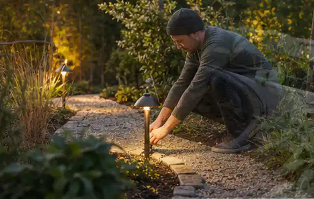 Person richtet eine niedrige warm leuchtende Gartenlampe am Kiesweg aus; Thema Familiengarten, Schwerpunkt Garten gestalten; Kontrolle.