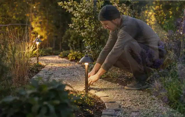 Person richtet eine niedrige warm leuchtende Gartenlampe am Kiesweg aus; Thema Familiengarten, Schwerpunkt Kosten; Ergebnis.