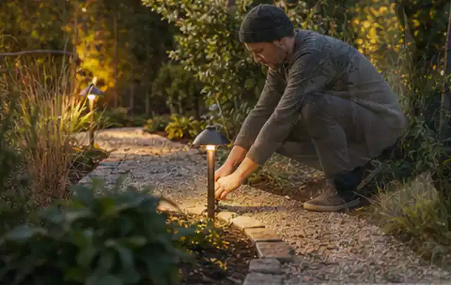 Person richtet eine niedrige warm leuchtende Gartenlampe am Kiesweg aus; Thema Familiengarten, Schwerpunkt Naturgarten; Praxisalltag.