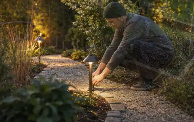 Person richtet eine niedrige warm leuchtende Gartenlampe am Kiesweg aus; Thema Familiengarten, Schwerpunkt Planung; Ausgangslage.