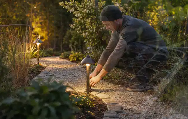 Person richtet eine niedrige warm leuchtende Gartenlampe am Kiesweg aus; Thema Familiengarten, Schwerpunkt Praxisbeispiele; Ergebnis.