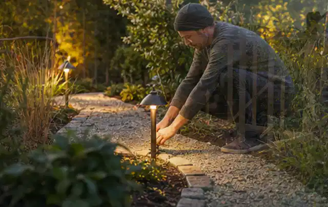 Person richtet eine niedrige warm leuchtende Gartenlampe am Kiesweg aus; Thema Familiengarten, Schwerpunkt Privatsphäre; Ausgangslage.