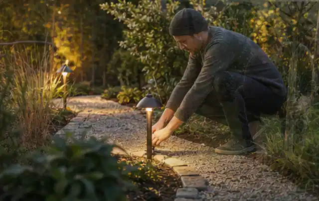 Person richtet eine niedrige warm leuchtende Gartenlampe am Kiesweg aus; Thema Familiengarten, Schwerpunkt Wegeführung; Kontrolle.