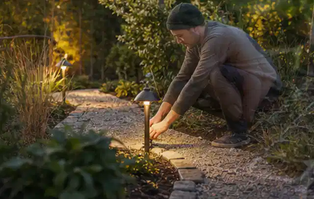 Person richtet eine niedrige warm leuchtende Gartenlampe am Kiesweg aus; Thema Feuerschalen, Schwerpunkt Checkliste; Ausgangslage.