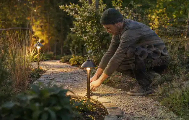 Person richtet eine niedrige warm leuchtende Gartenlampe am Kiesweg aus; Thema Feuerschalen, Schwerpunkt Ersatzteile; Kontrolle.