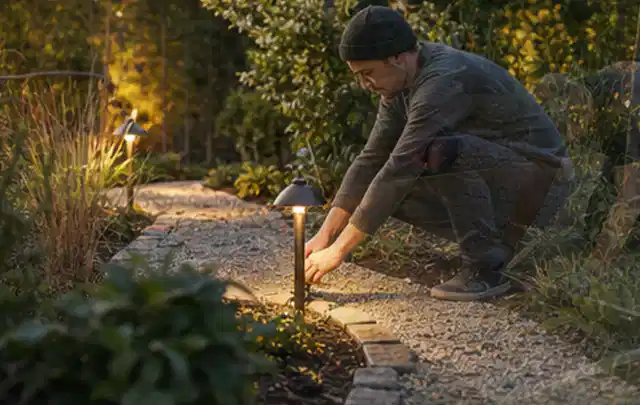 Person richtet eine niedrige warm leuchtende Gartenlampe am Kiesweg aus; Thema Feuerschalen, Schwerpunkt Gärten mit großer Fläche; Umsetzung.