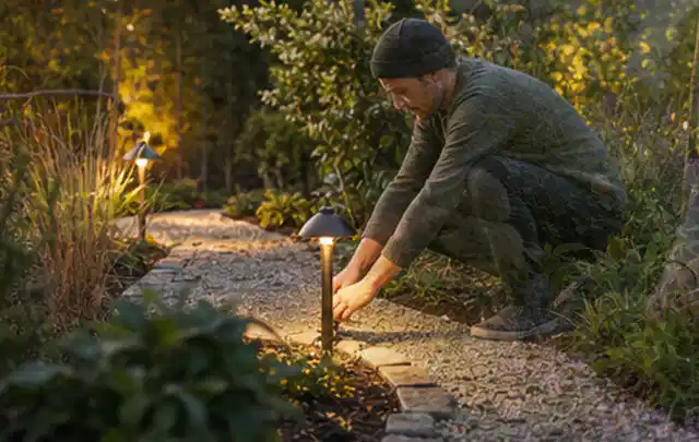 Person richtet eine niedrige warm leuchtende Gartenlampe am Kiesweg aus; Thema Feuerschalen, Schwerpunkt Reinigung; Umsetzung.