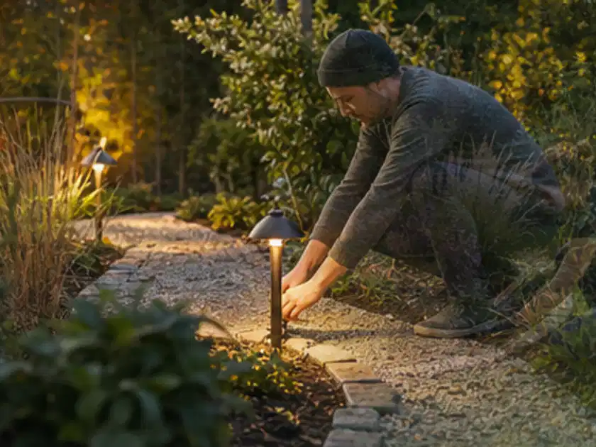 Person richtet eine niedrige warm leuchtende Gartenlampe am Kiesweg aus; Thema Feuerschalen, Schwerpunkt Sicherheit; Titelbild.