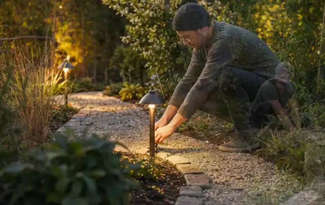 Person richtet eine niedrige warm leuchtende Gartenlampe am Kiesweg aus; Thema Feuerschalen, Schwerpunkt Vergleich; Kontrolle.