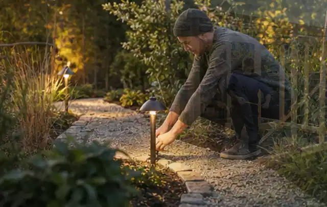 Person richtet eine niedrige warm leuchtende Gartenlampe am Kiesweg aus; Thema Feuerschalen, Schwerpunkt Winterlager; Umsetzung.