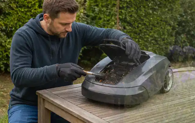 Person wartet einen geöffneten Mähroboter auf einem stabilen Gartentisch; Thema Freischneider, Schwerpunkt Checkliste; Kontrolle.