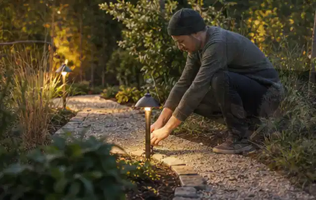 Person richtet eine niedrige warm leuchtende Gartenlampe am Kiesweg aus; Thema Gabionen, Schwerpunkt Aufbau; Praxisalltag.