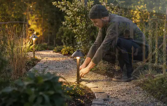 Person richtet eine niedrige warm leuchtende Gartenlampe am Kiesweg aus; Thema Gabionen, Schwerpunkt Baurecht; Praxisalltag.