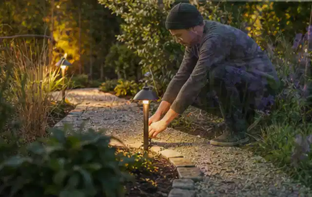 Person richtet eine niedrige warm leuchtende Gartenlampe am Kiesweg aus; Thema Gabionen, Schwerpunkt Dimensionierung; Kontrolle.
