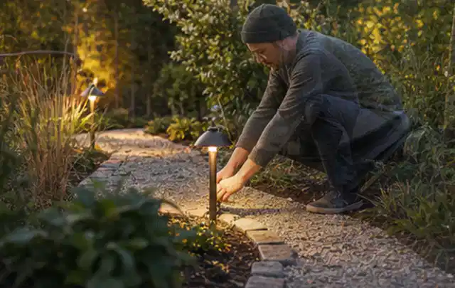 Person richtet eine niedrige warm leuchtende Gartenlampe am Kiesweg aus; Thema Gabionen, Schwerpunkt Materialwahl; Ergebnis.