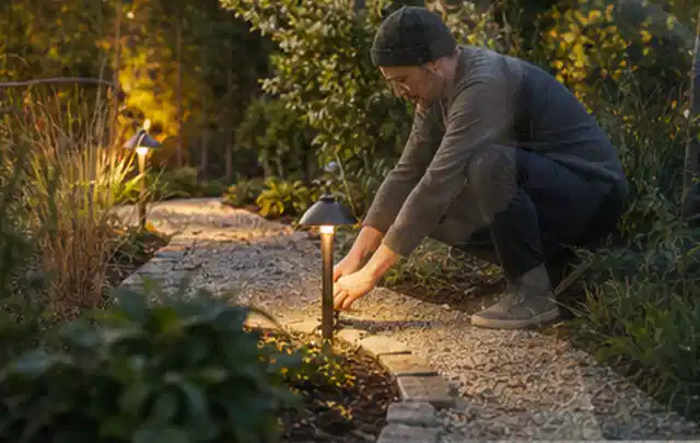 Person richtet eine niedrige warm leuchtende Gartenlampe am Kiesweg aus; Thema Gabionen, Schwerpunkt Nachhaltigkeit; Ergebnis.