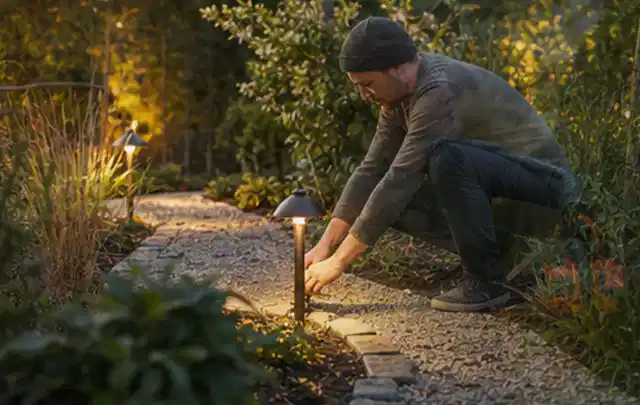 Person richtet eine niedrige warm leuchtende Gartenlampe am Kiesweg aus; Thema Gardena Sileno, Schwerpunkt Anwendung; Ausgangslage.