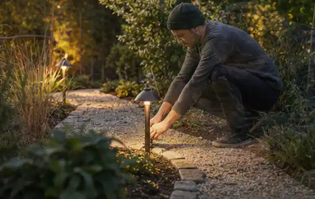 Person richtet eine niedrige warm leuchtende Gartenlampe am Kiesweg aus; Thema Gardena Sileno, Schwerpunkt Bestenliste; Umsetzung.