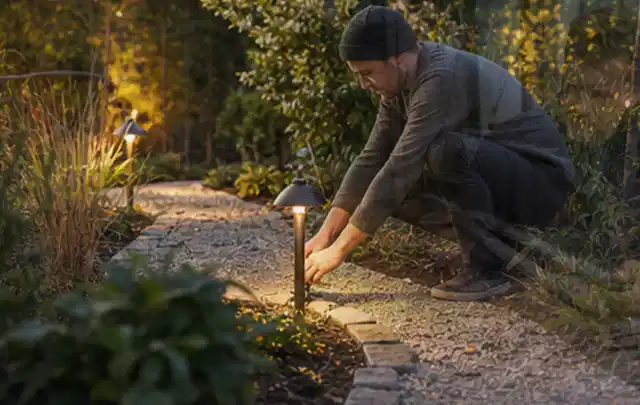 Person richtet eine niedrige warm leuchtende Gartenlampe am Kiesweg aus; Thema Gardena Sileno, Schwerpunkt Ersatzteile; Ergebnis.