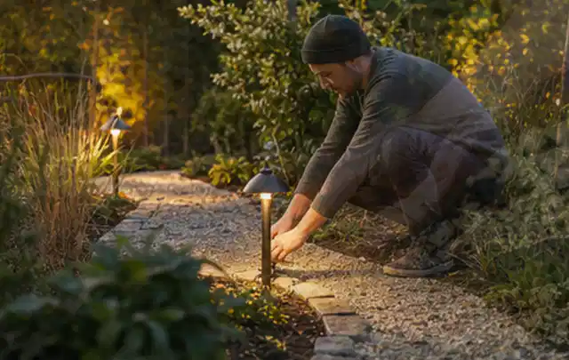 Person richtet eine niedrige warm leuchtende Gartenlampe am Kiesweg aus; Thema Gardena Sileno, Schwerpunkt Gärten mit großer Fläche; Praxisalltag.