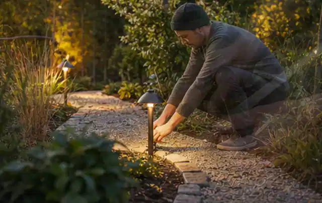Person richtet eine niedrige warm leuchtende Gartenlampe am Kiesweg aus; Thema Gardena Sileno, Schwerpunkt Leistung; Umsetzung.