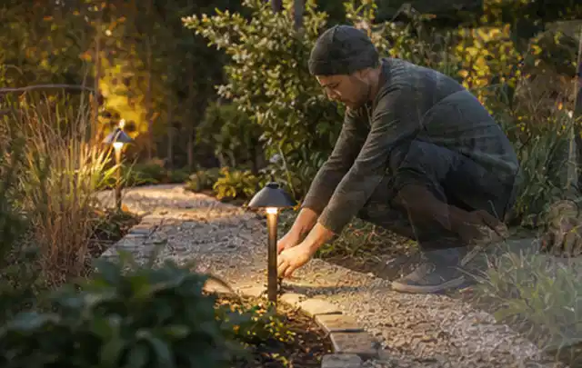 Person richtet eine niedrige warm leuchtende Gartenlampe am Kiesweg aus; Thema Gardena Sileno, Schwerpunkt Mähroboter; Umsetzung.