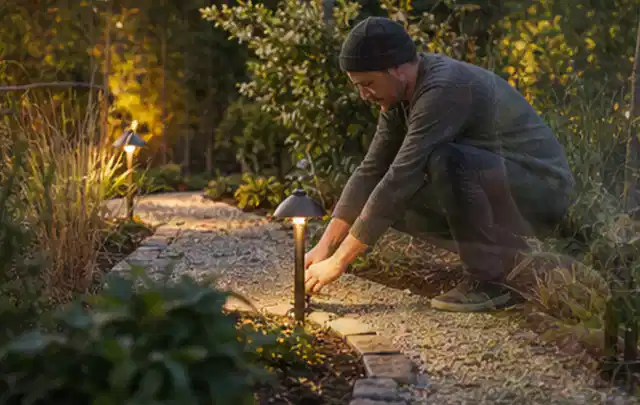Person richtet eine niedrige warm leuchtende Gartenlampe am Kiesweg aus; Thema Gardena Sileno, Schwerpunkt Wartung; Umsetzung.