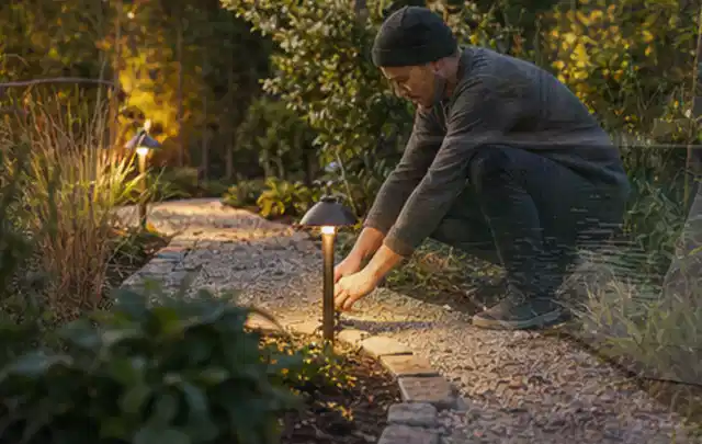 Person richtet eine niedrige warm leuchtende Gartenlampe am Kiesweg aus; Thema Garten am Neubau, Schwerpunkt Anleitung; Ausgangslage.