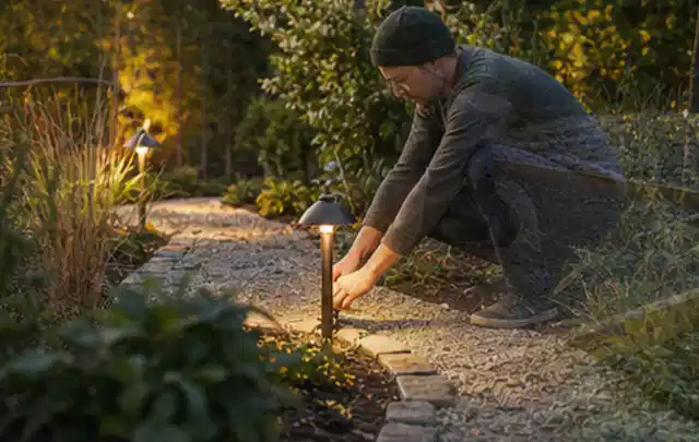 Person richtet eine niedrige warm leuchtende Gartenlampe am Kiesweg aus; Thema Garten am Neubau, Schwerpunkt Checkliste; Praxisalltag.