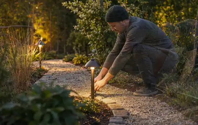 Person richtet eine niedrige warm leuchtende Gartenlampe am Kiesweg aus; Thema Garten am Neubau, Schwerpunkt Fehlerhilfe; Ergebnis.
