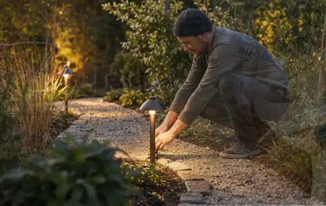 Person richtet eine niedrige warm leuchtende Gartenlampe am Kiesweg aus; Thema Garten am Neubau, Schwerpunkt Naturgarten; Ergebnis.