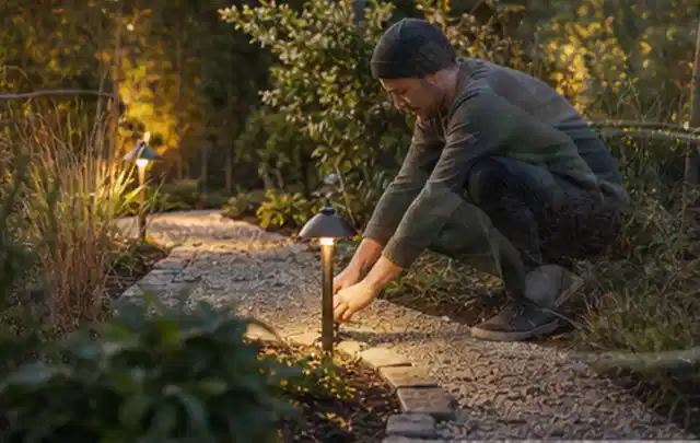 Person richtet eine niedrige warm leuchtende Gartenlampe am Kiesweg aus; Thema Garten am Neubau, Schwerpunkt Pflegeleichtigkeit; Kontrolle.