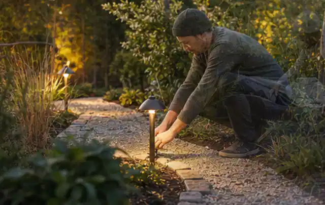 Person richtet eine niedrige warm leuchtende Gartenlampe am Kiesweg aus; Thema Garten gestalten, Schwerpunkt Themenwelt; Ergebnis.