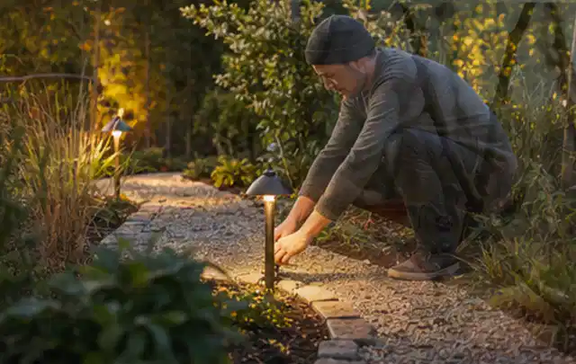 Person richtet eine niedrige warm leuchtende Gartenlampe am Kiesweg aus; Thema Gartenhaus Aufbau, Schwerpunkt Fehlerhilfe; Umsetzung.