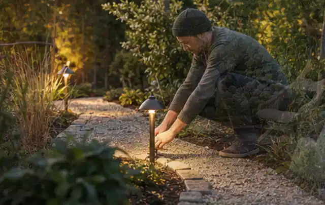 Person richtet eine niedrige warm leuchtende Gartenlampe am Kiesweg aus; Thema Gartenhaus Aufbau, Schwerpunkt Materialwahl; Praxisalltag.