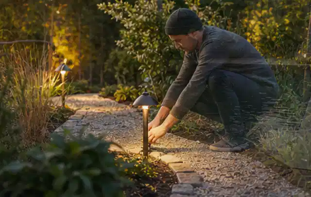 Person richtet eine niedrige warm leuchtende Gartenlampe am Kiesweg aus; Thema Gartenhaus Aufbau, Schwerpunkt Planung; Ergebnis.