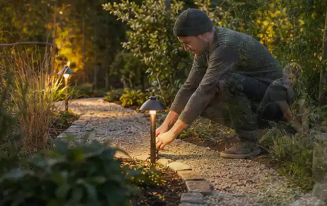 Person richtet eine niedrige warm leuchtende Gartenlampe am Kiesweg aus; Thema Gartenhaus aus Holz, Schwerpunkt Fundament; Umsetzung.