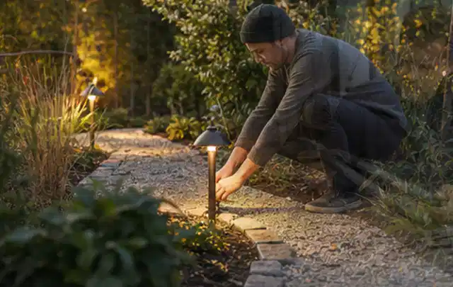 Person richtet eine niedrige warm leuchtende Gartenlampe am Kiesweg aus; Thema Gartenhaus aus Holz, Schwerpunkt Gärten mit kleiner Fläche; Umsetzung.