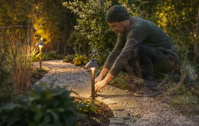 Person richtet eine niedrige warm leuchtende Gartenlampe am Kiesweg aus; Thema Gartenhaus aus Holz, Schwerpunkt Gestaltung; Ergebnis.