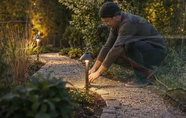 Person richtet eine niedrige warm leuchtende Gartenlampe am Kiesweg aus; Thema Gartenhaus aus Holz, Schwerpunkt Kosten; Kontrolle.