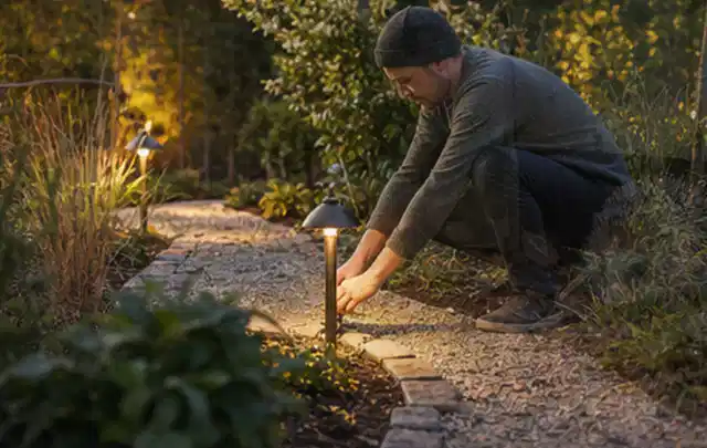 Person richtet eine niedrige warm leuchtende Gartenlampe am Kiesweg aus; Thema Gartenhaus aus Holz, Schwerpunkt Materialwahl; Praxisalltag.