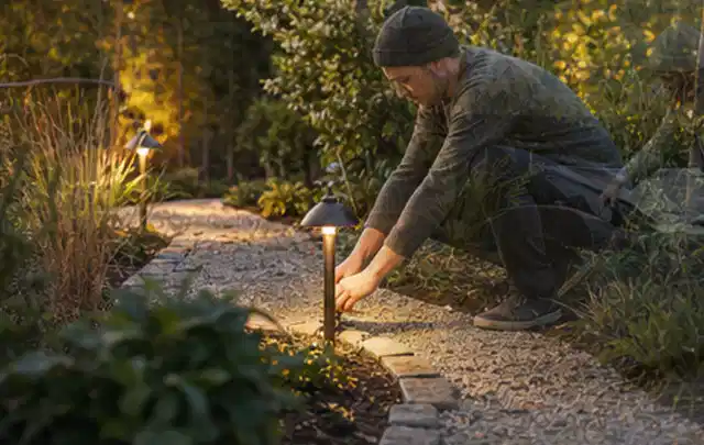 Person richtet eine niedrige warm leuchtende Gartenlampe am Kiesweg aus; Thema Gartenhaus aus Holz, Schwerpunkt Pflege; Praxisalltag.