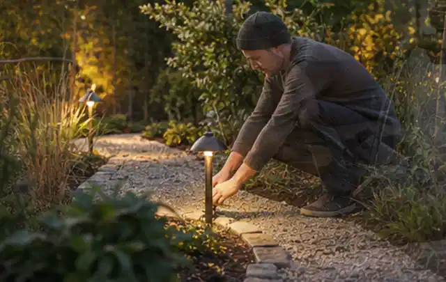 Person richtet eine niedrige warm leuchtende Gartenlampe am Kiesweg aus; Thema Gartenhaus aus Holz, Schwerpunkt Planung; Praxisalltag.