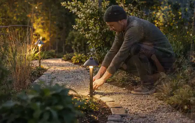 Person richtet eine niedrige warm leuchtende Gartenlampe am Kiesweg aus; Thema Gartenhaus aus Kunststoff, Schwerpunkt Baurecht; Praxisalltag.