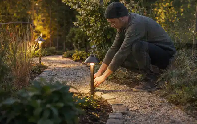 Person richtet eine niedrige warm leuchtende Gartenlampe am Kiesweg aus; Thema Gartenhaus aus Kunststoff, Schwerpunkt Fundament; Ausgangslage.