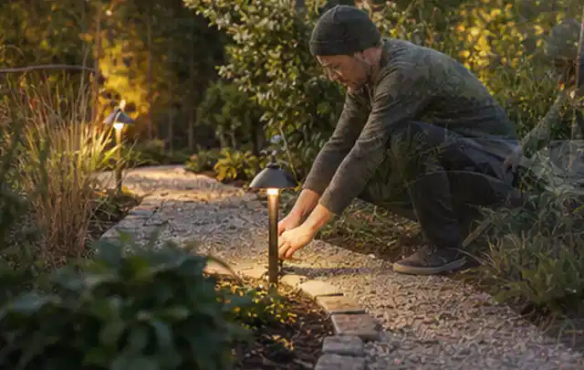 Person richtet eine niedrige warm leuchtende Gartenlampe am Kiesweg aus; Thema Gartenhaus aus Kunststoff, Schwerpunkt Gestaltung; Praxisalltag.